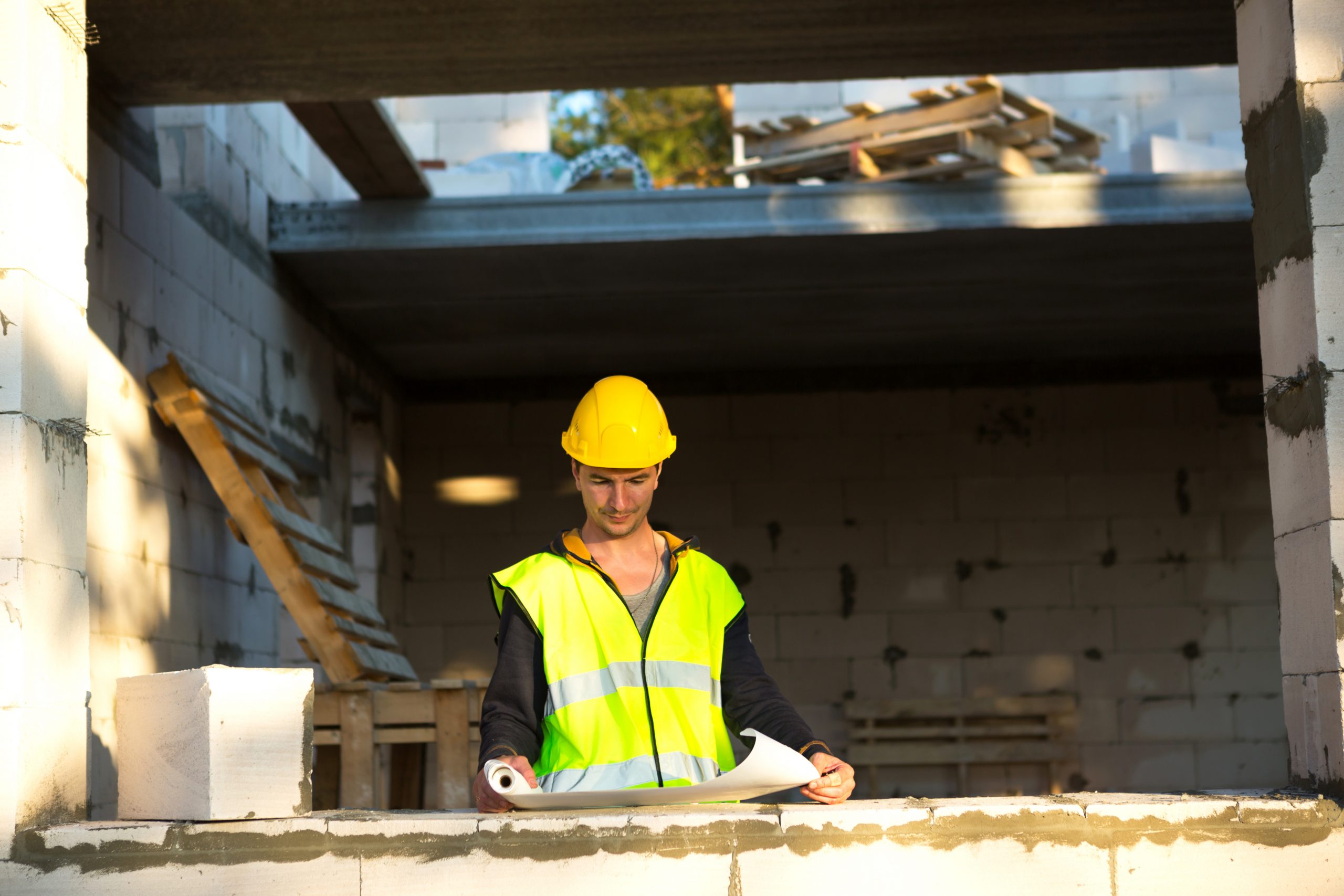 A construction engineer and designer in a yellow hardhat studies a drawing of a building on a construction site. A house made of porous concrete blocks, a worker in the process at the window and wall