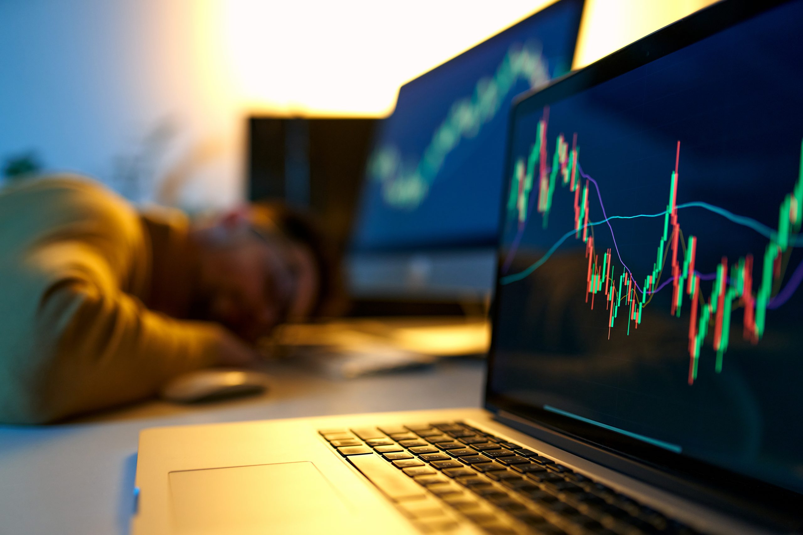 Fatigue man sleeps on his home office desk while trading, browsing online stock investments at night. Tired novice male trader sleeping near monitor with stock chart at workplace.