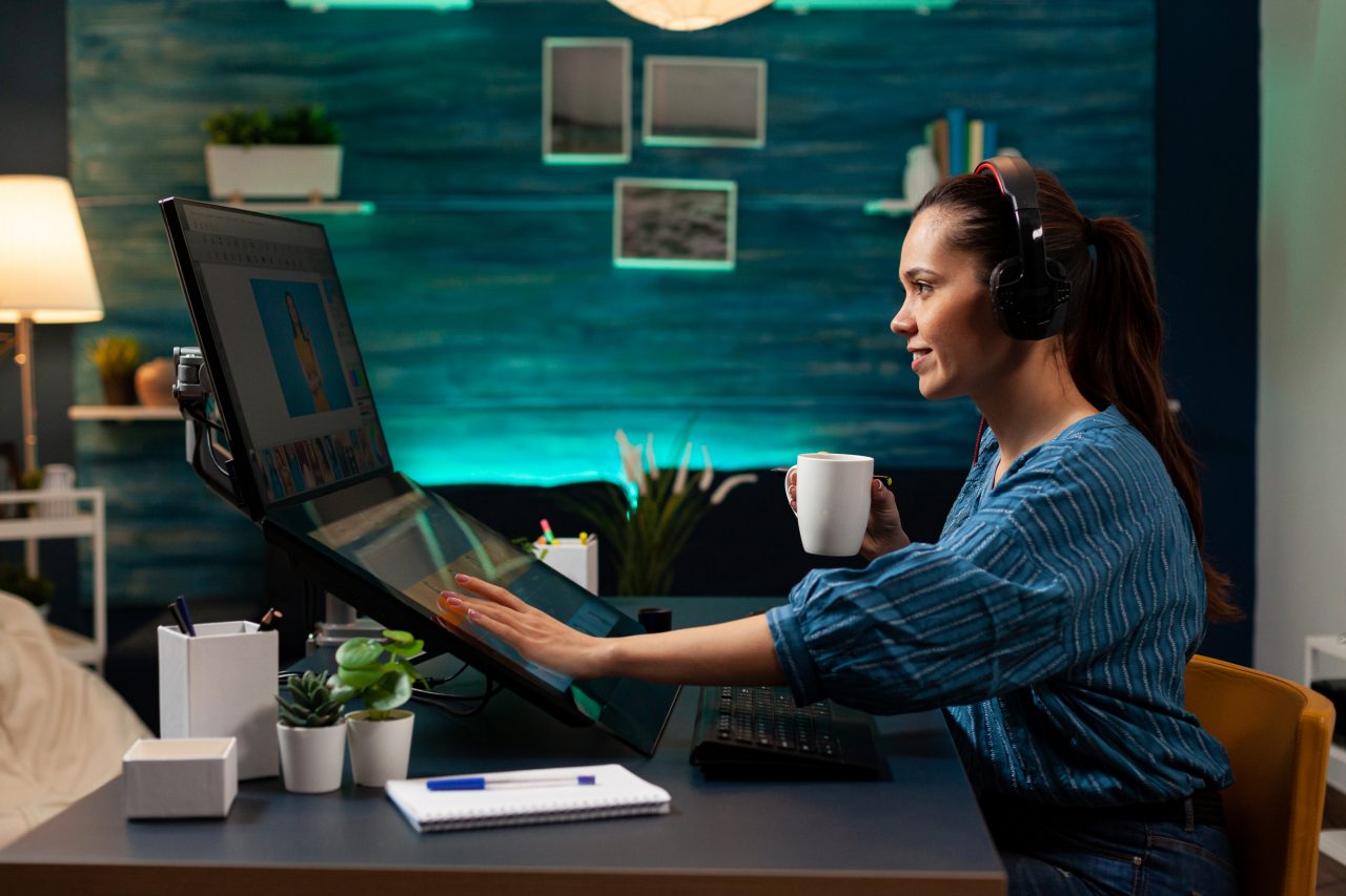 Graphic software editor with headphones working on photography project using touchpad computer monitor sceen display. Woman at workplace doing photo retouching with modern interface