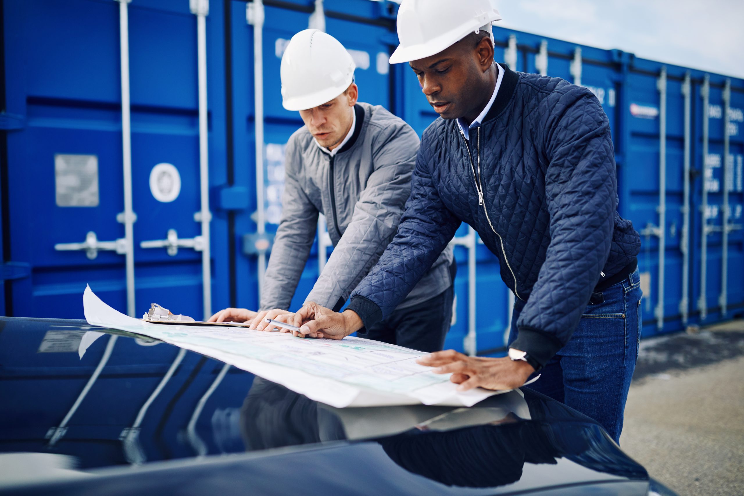 Two engineers leaning on the hood of a truck discussing blueprints while standing by freight containers on a commercial shipping dock